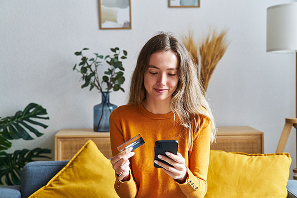 Smiling woman holds credit card and cell phone while sitting on couch.